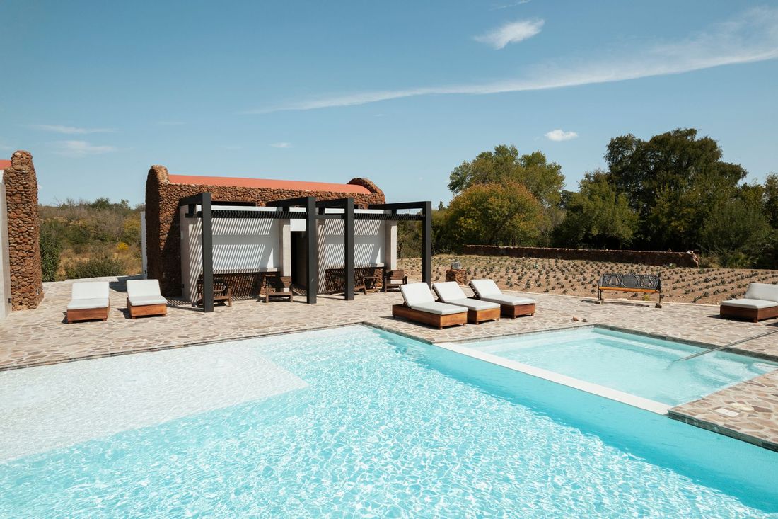 Escena junto a la piscina con tumbonas y un edificio con pérgola. Día soleado.