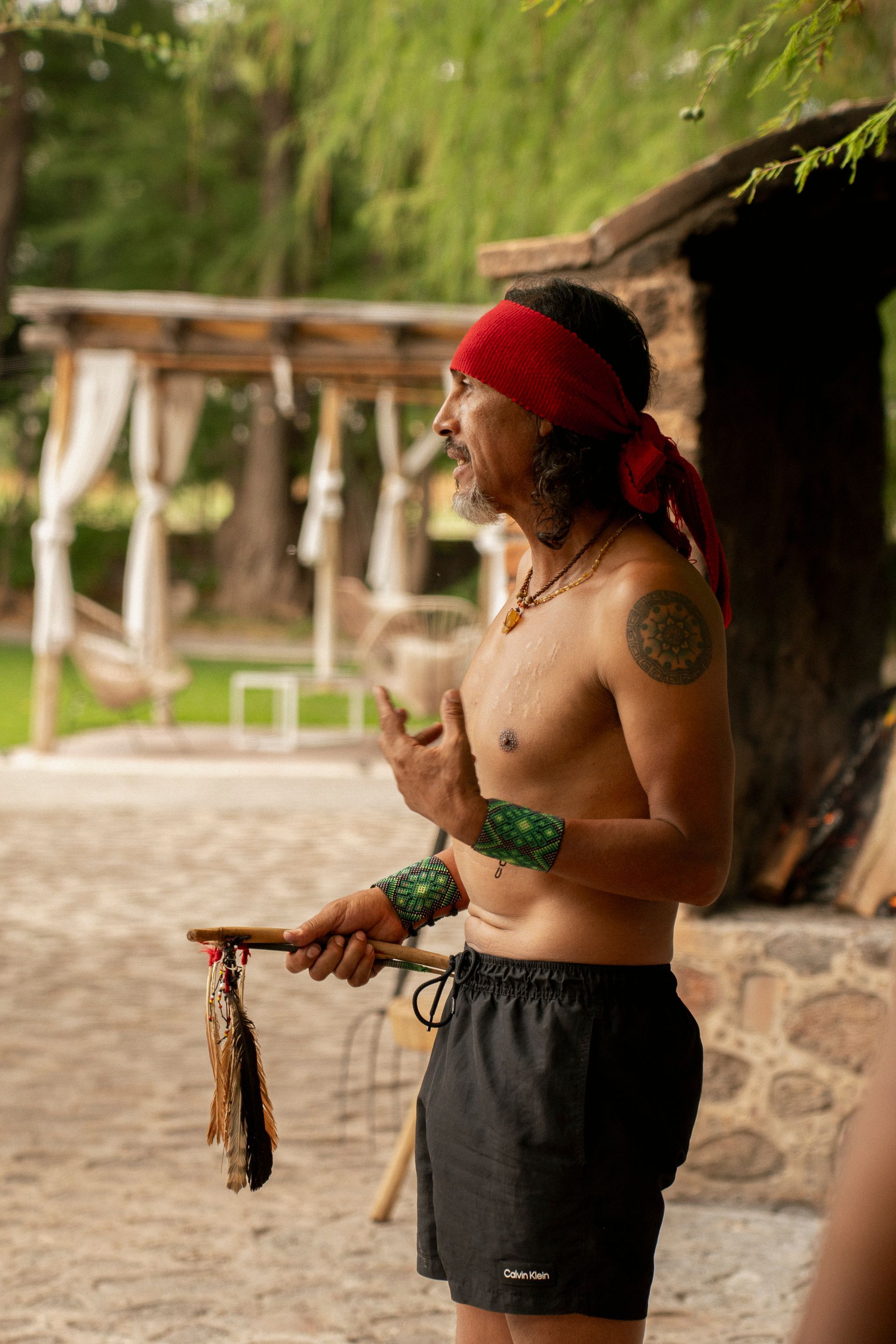 Hombre con cinta roja en la cabeza, sin camisa, gesticulando. Al aire libre, sosteniendo un palo con plumas.