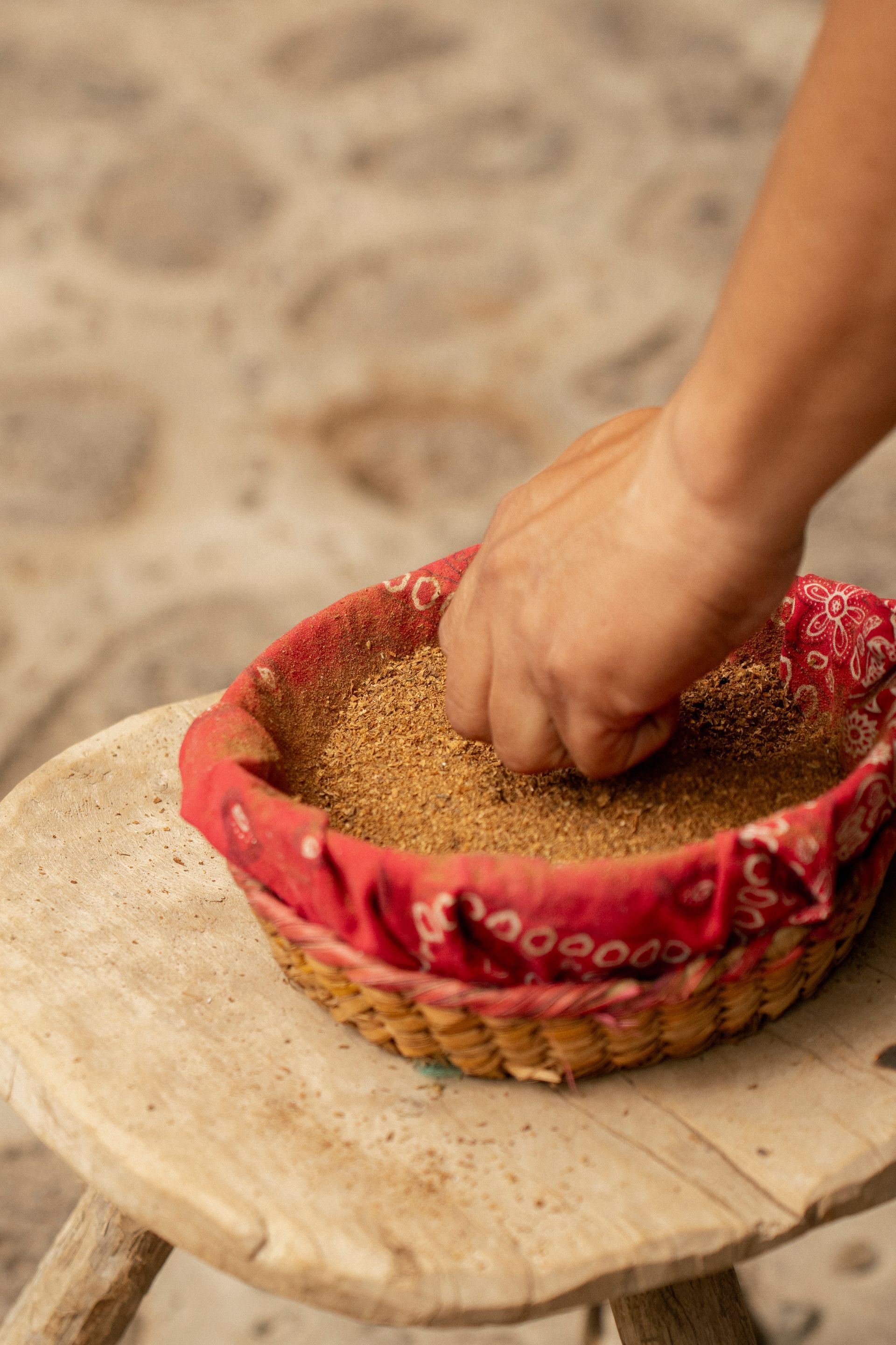 Removiendo granos a mano en una cesta forrada de rojo, colocada sobre un taburete de madera, al aire libre.