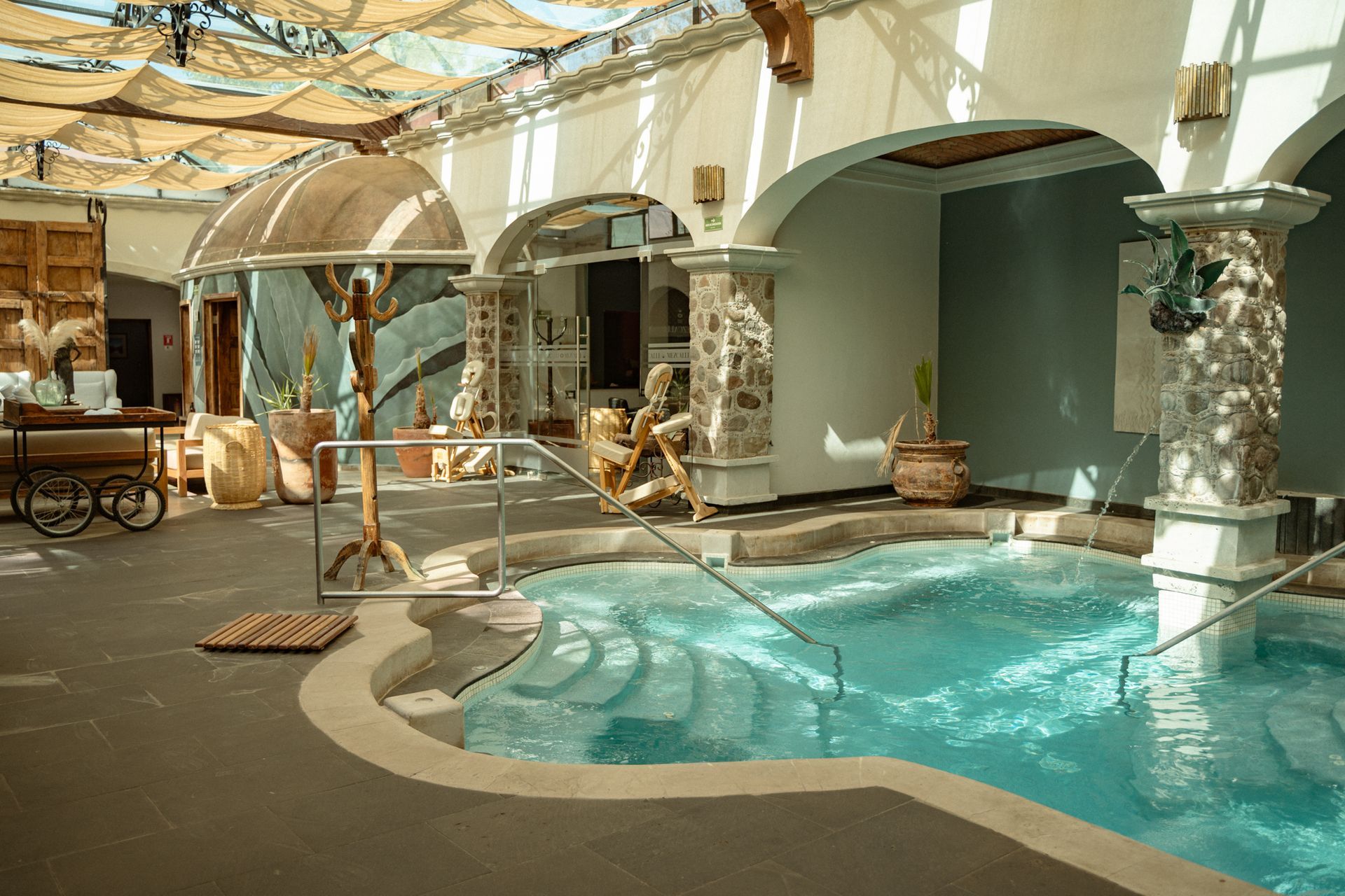 Piscina de hidromasaje interior con columnas y arcos de piedra, bajo un toldo que filtra la luz.