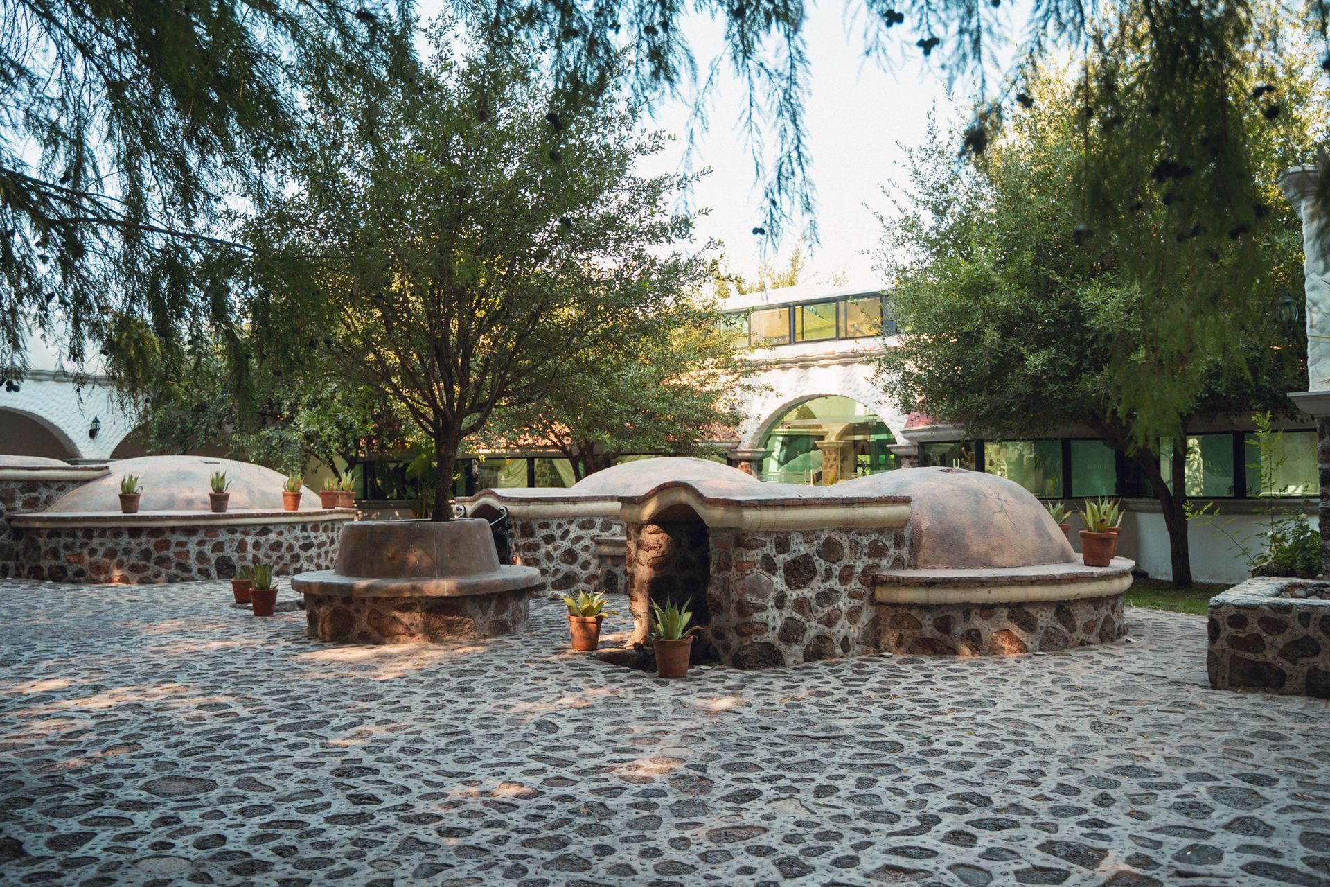 Patio con estructuras de piedra y entradas arqueadas; árboles en lo alto y un edificio con ventanas al fondo.