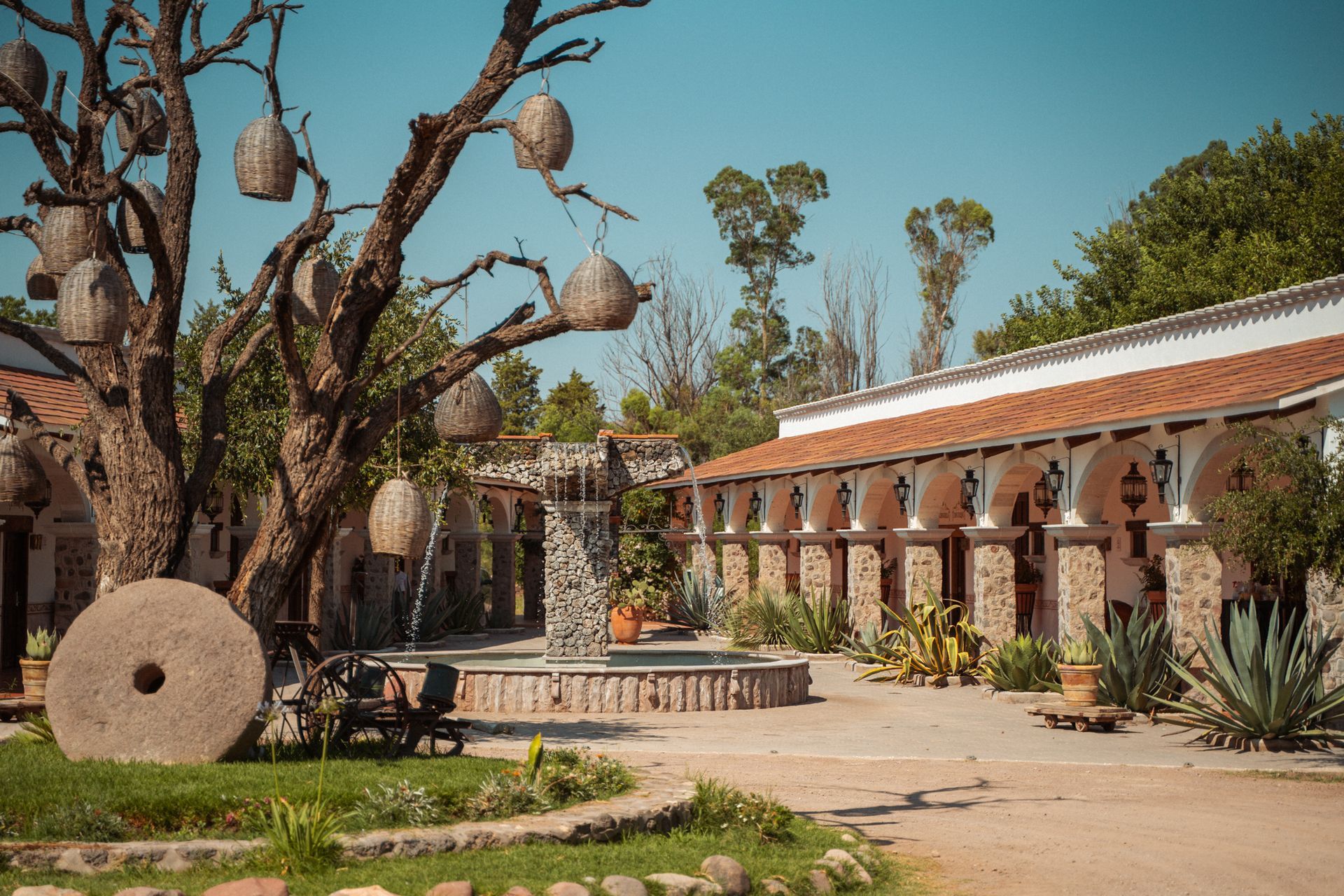 Patio con fuente, piedra de molino y un árbol con adornos colgantes junto a un edificio de estilo español.