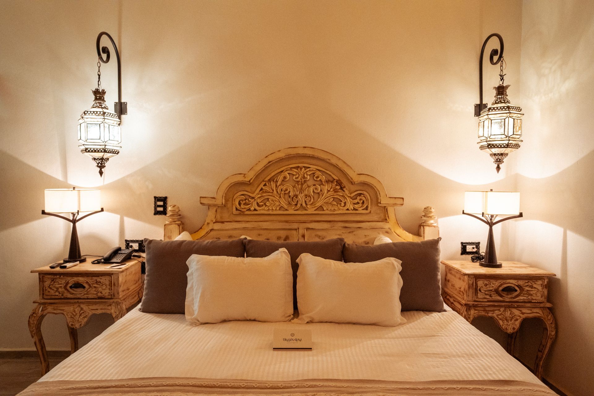 Dormitorio con cabecero ornamentado, mesitas de noche y faroles. La cama tiene almohadas y manta. Iluminación tenue.