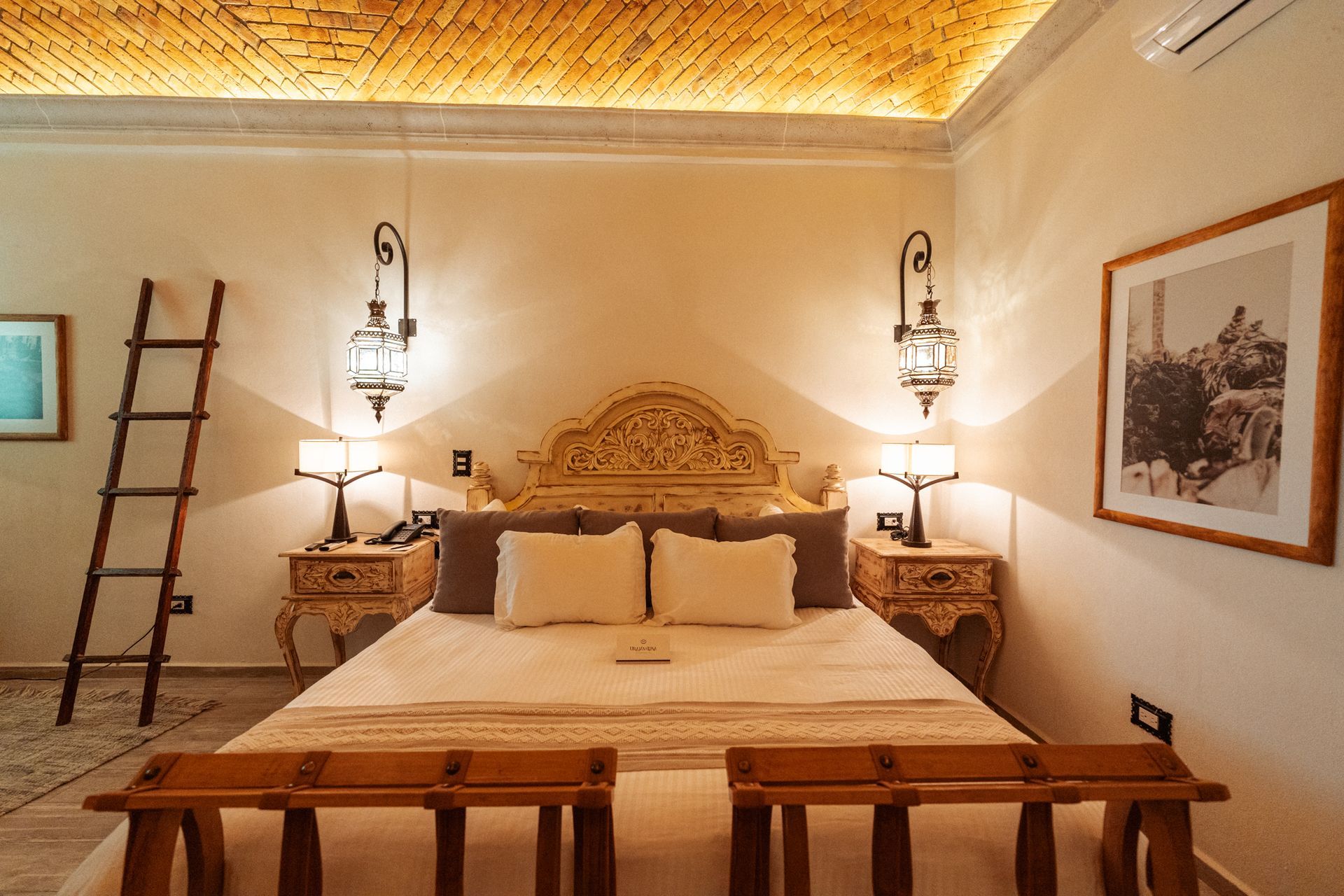 Dormitorio con cabecero ornamentado, dos mesitas de noche con lámparas, faroles colgantes y obras de arte. 