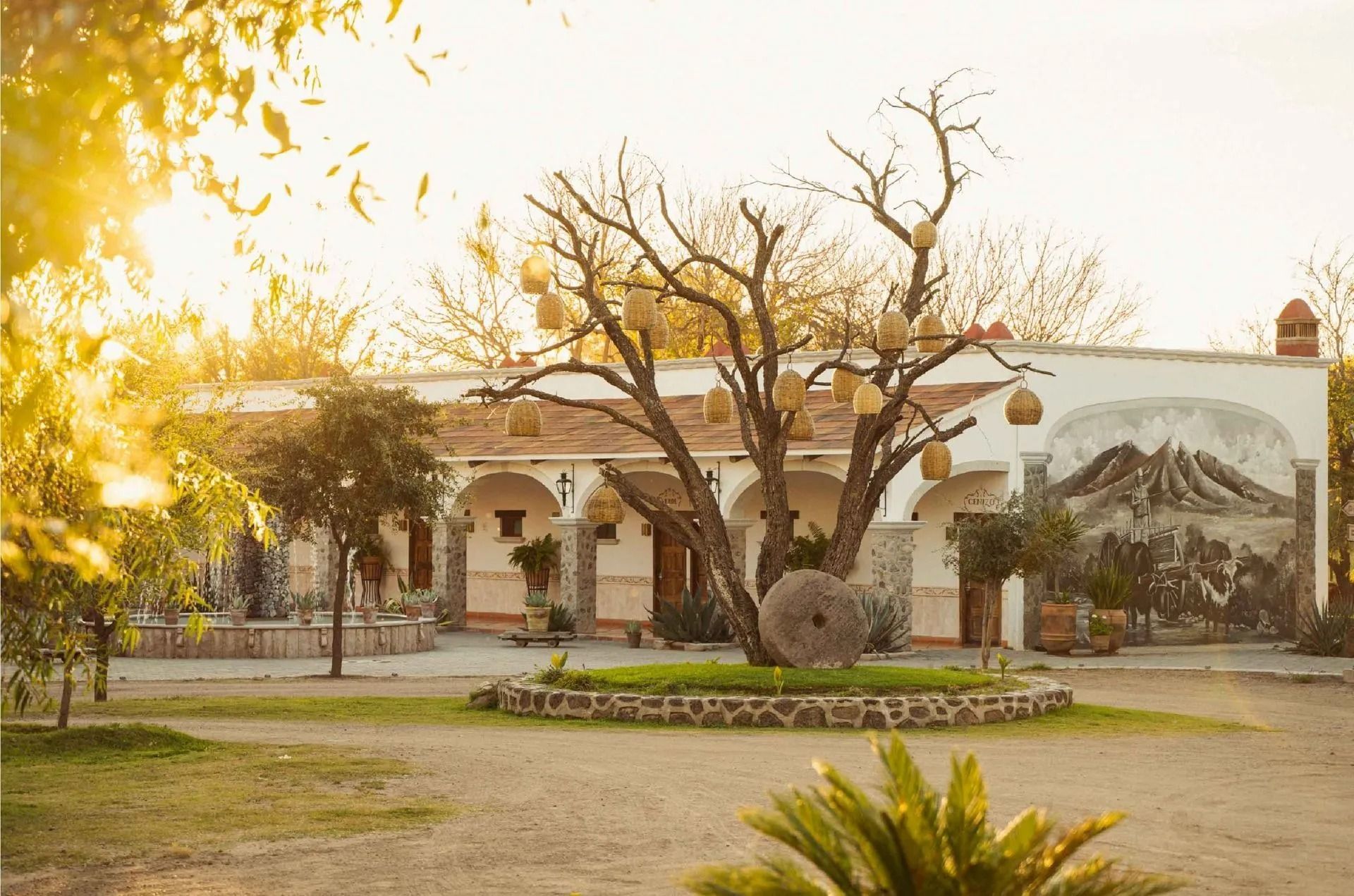 Hacienda de una sola planta, bañada por el sol, con pasillos arqueados, un árbol central adornado con faroles y un mural