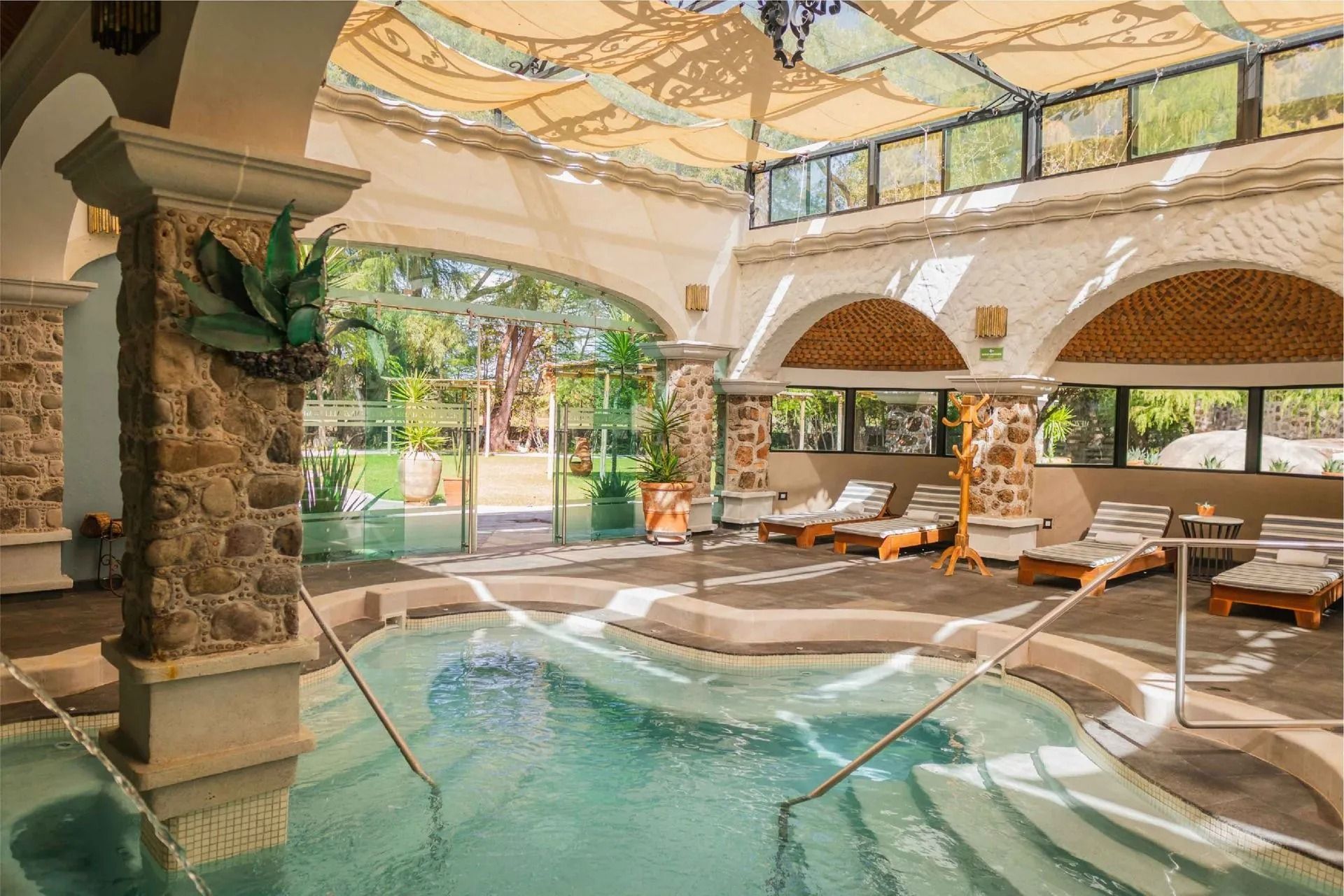 Zona de piscina cubierta con columnas de piedra, techos abovedados y tumbonas, rodeada de ventanales y  vegetación.