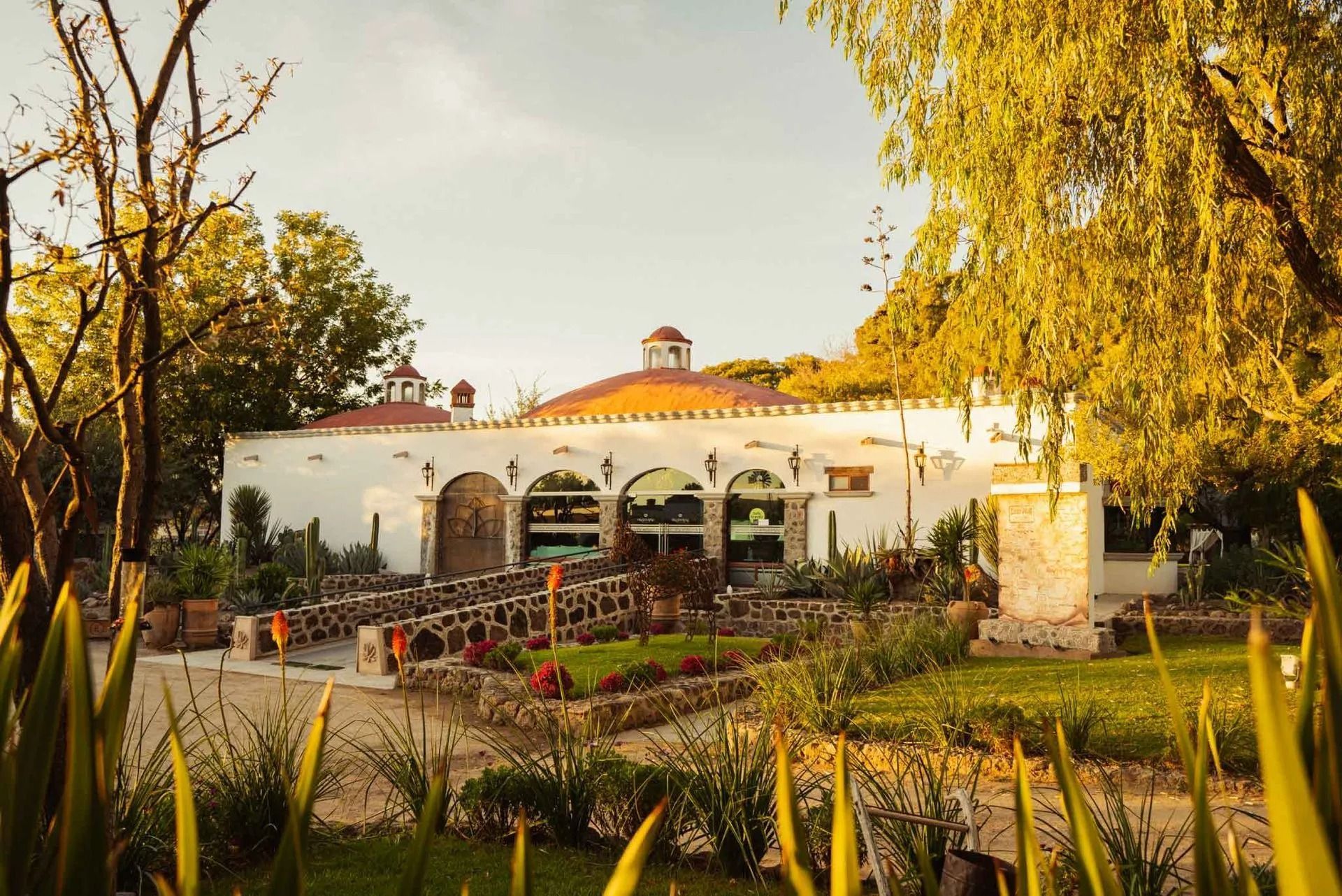 Hacienda blanca bañada por el sol, con puertas arqueadas, puentes de piedra y  jardines bajo un cielo despejado.