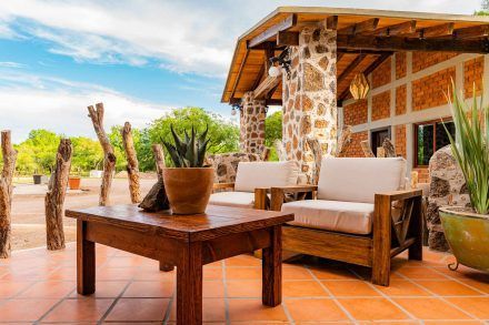 Outdoor patio with wood furniture, stone columns, terracotta tiles, and potted plants under a sunny sky.