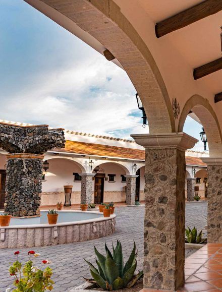 Courtyard with arches, stone columns, and terracotta pots. Fountain, blue sky.