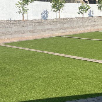 Green artificial turf lawn with concrete pathways and a tiered wall with small trees.