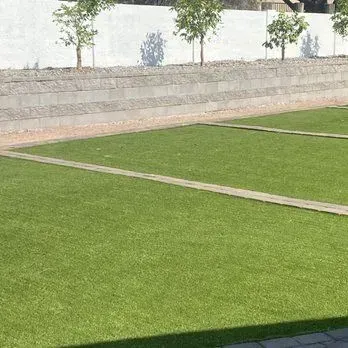 Green artificial turf lawn with concrete pathways and a tiered wall with small trees.