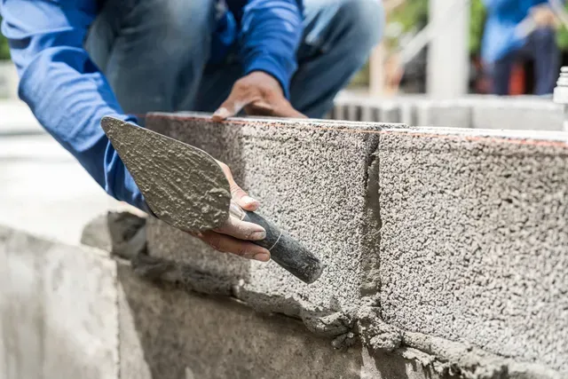 A construction worker applying mortar to a cinder block wall with a trowel outdoors.
