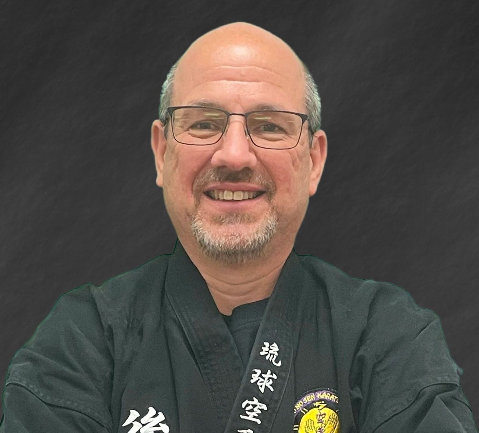 A man wearing glasses and a black karate uniform is smiling for the camera.