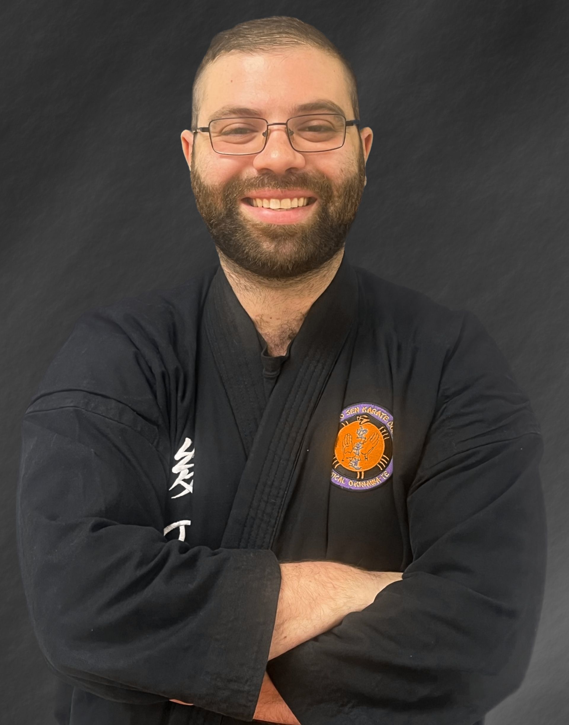 A man in a black karate uniform is smiling with his arms crossed.