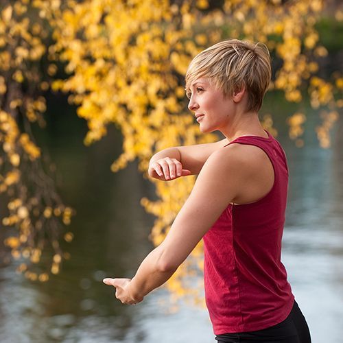 A woman in a red tank top is stretching her arms in front of a body of water.