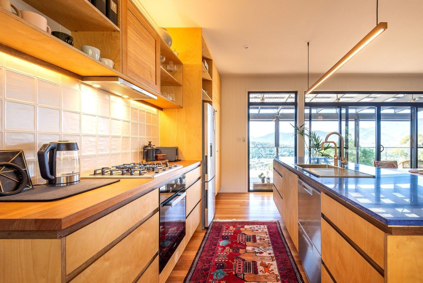 A brightly lit kitchen with brown cabinets, brown wooden floor, and a red carpet. 