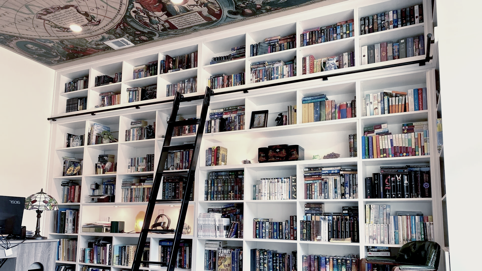 White bookcase filled with books, with a rolling ladder in front of it and a map on the ceiling.