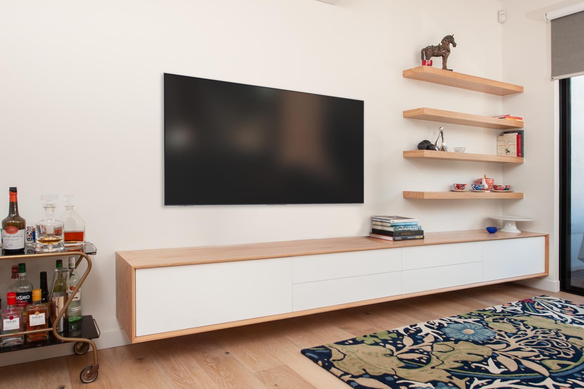 Modern living room with a mounted TV, floating shelves, and a bar cart; rug on a wooden floor.