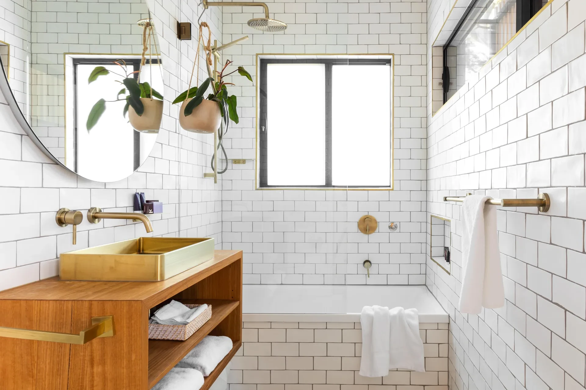 A bathroom with white tiles, a tub, and brown wooden cabinet below a golden sink. 