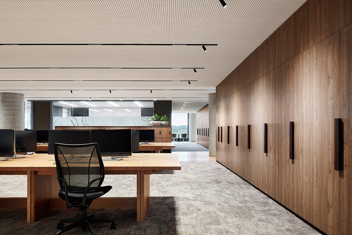Modern open-plan office with wooden desks, mesh chairs, wood-paneled wall, and patterned ceiling.