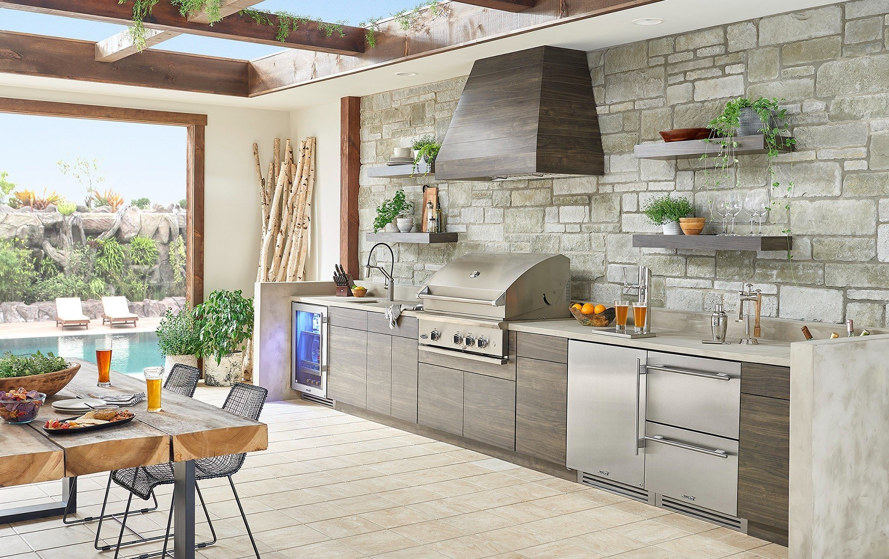 Outdoor kitchen with grill, sink, and stone wall, with pool in the background.