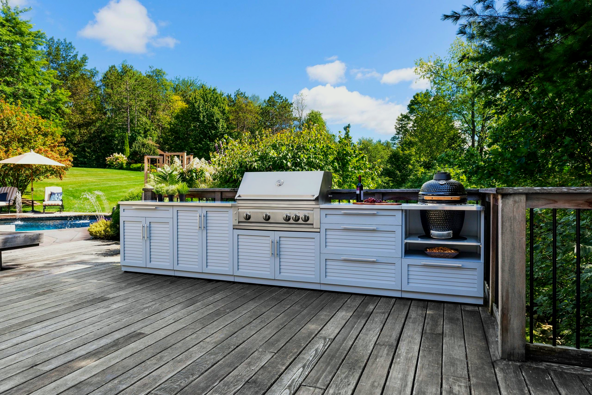 Outdoor kitchen with grill on a wooden deck overlooking a yard, blue sky above.