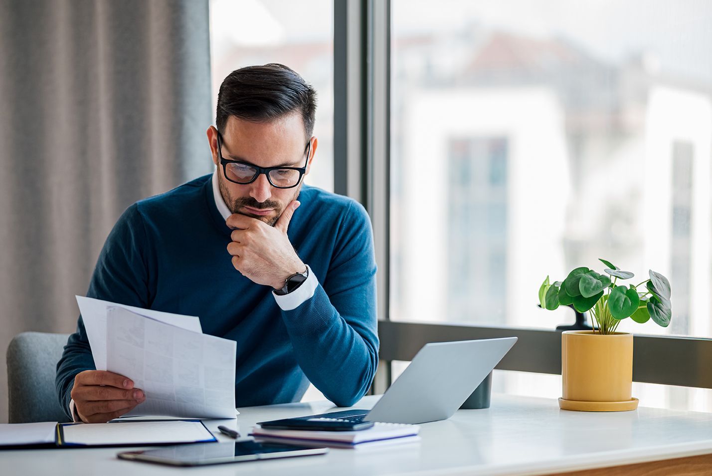 Une personne concentrée, vêtue d'un pull bleu, est assise à un bureau avec un ordinateur portable