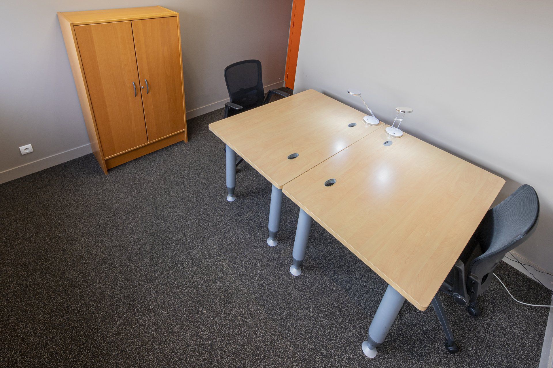 Espace de bureau avec deux bureaux, des chaises et une armoire en bois. Moquette grise, murs gris, touches d'orange.