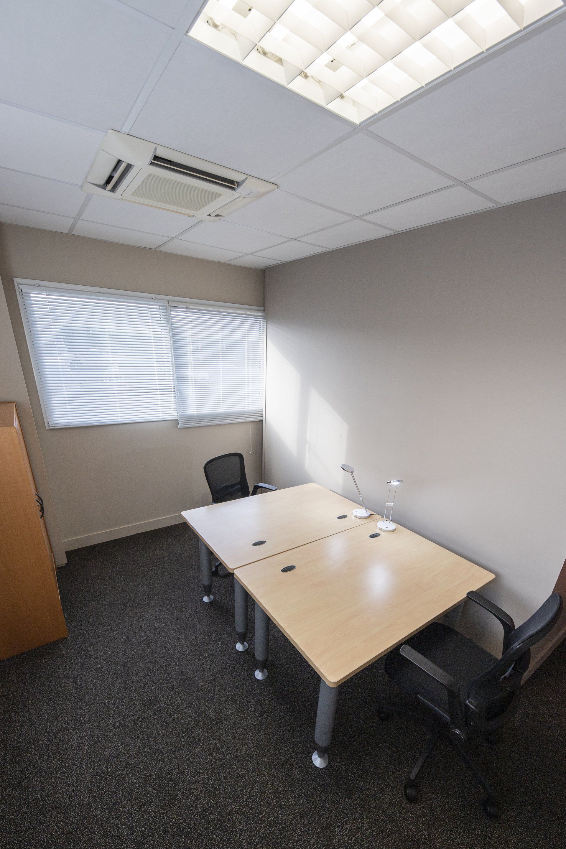 Petit bureau avec deux bureaux, des chaises, une fenêtre avec stores et des néons au plafond.
