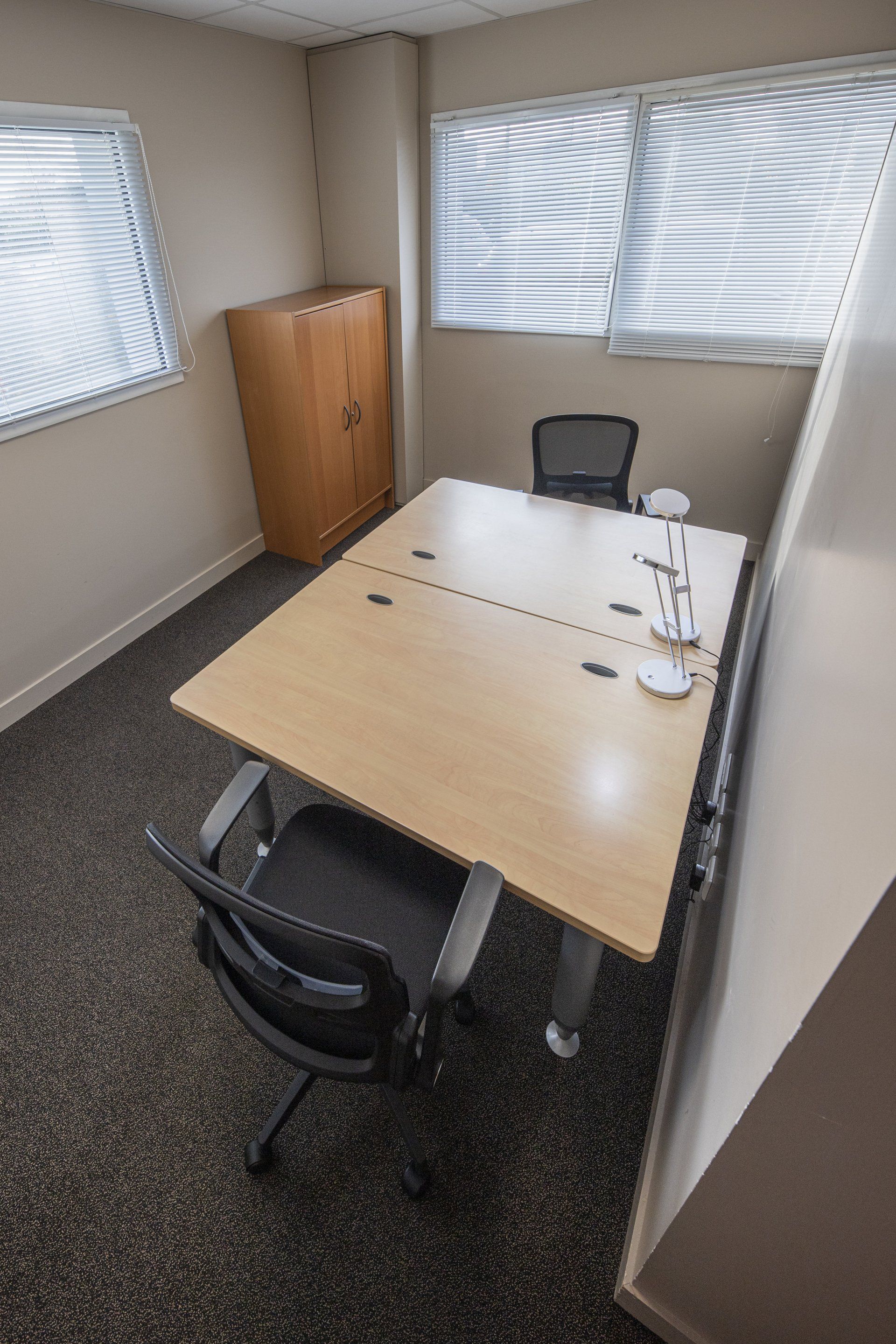 Petit bureau avec une table rectangulaire, deux chaises, une armoire et deux fenêtres avec stores.