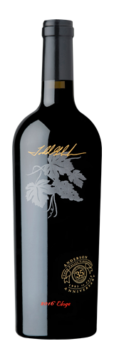 A black wine bottle with gold cursive script, an etched grape vine graphic, and a round anniversary seal on the label.