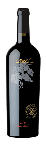 A dark wine bottle with a gold signature, a silver grape leaf illustration, and a circular emblem label near the base.