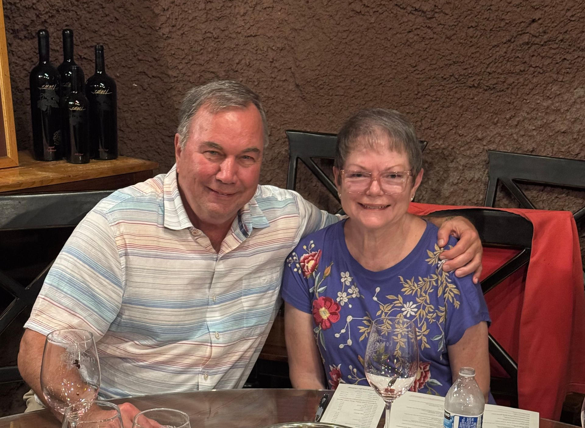 A smiling man and woman sit together at a table, likely in a restaurant with wine bottles visible in the background.