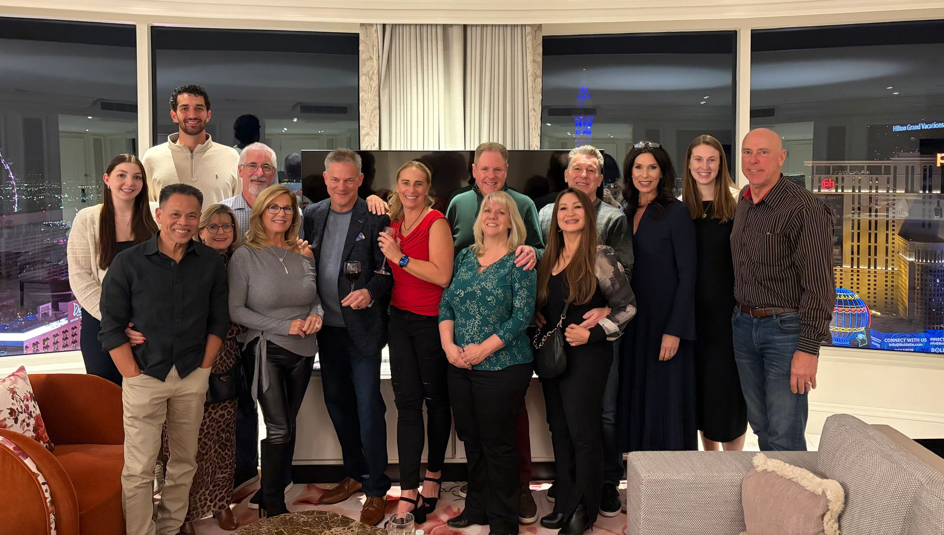 A group of fourteen people stand smiling and posing for a photo in a brightly lit room with large windows at night.