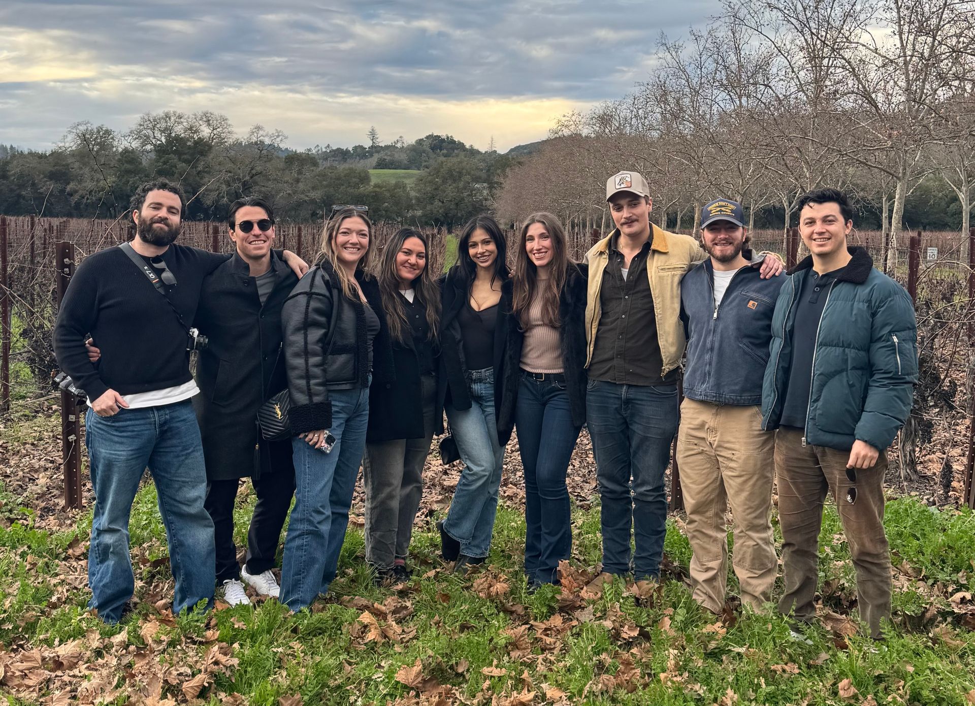 A group of nine friends smiling while standing together in a vineyard during the late afternoon.
