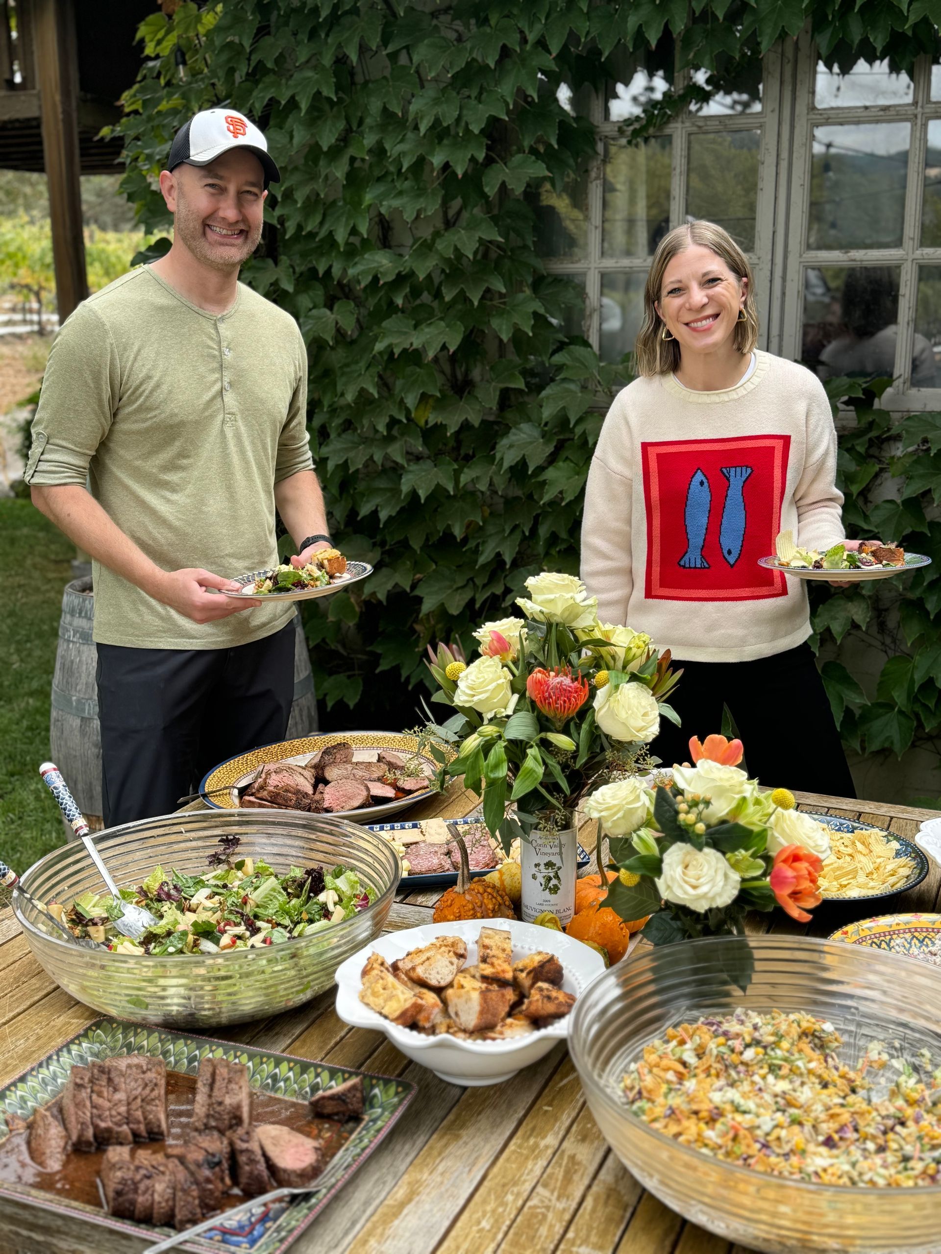 A smiling man and woman standing behind an outdoor table spread with various dishes, salads, and flower centerpieces.