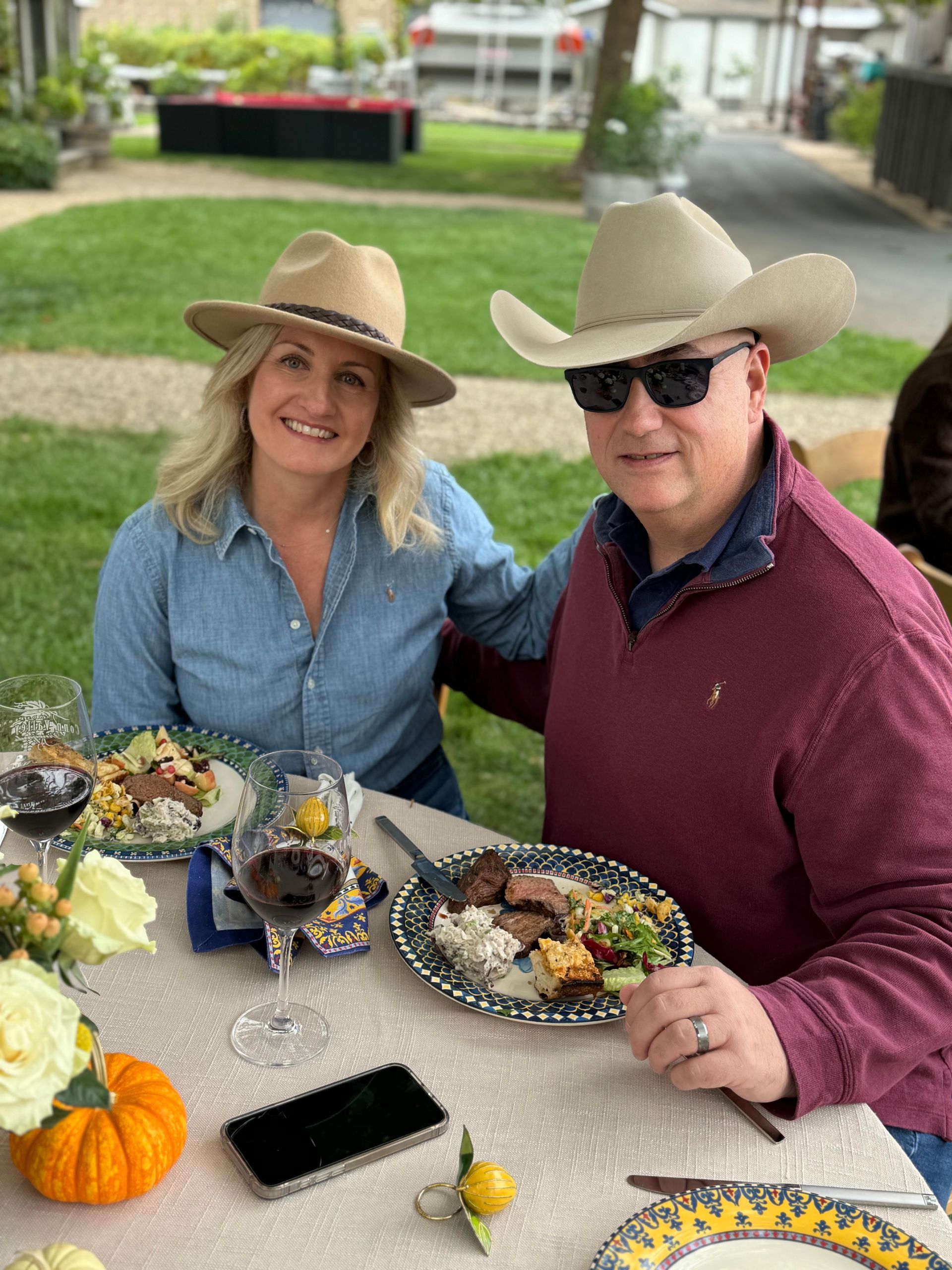 A smiling couple in hats dining outdoors at a table set with wine, food, and small pumpkins on a sunny day.