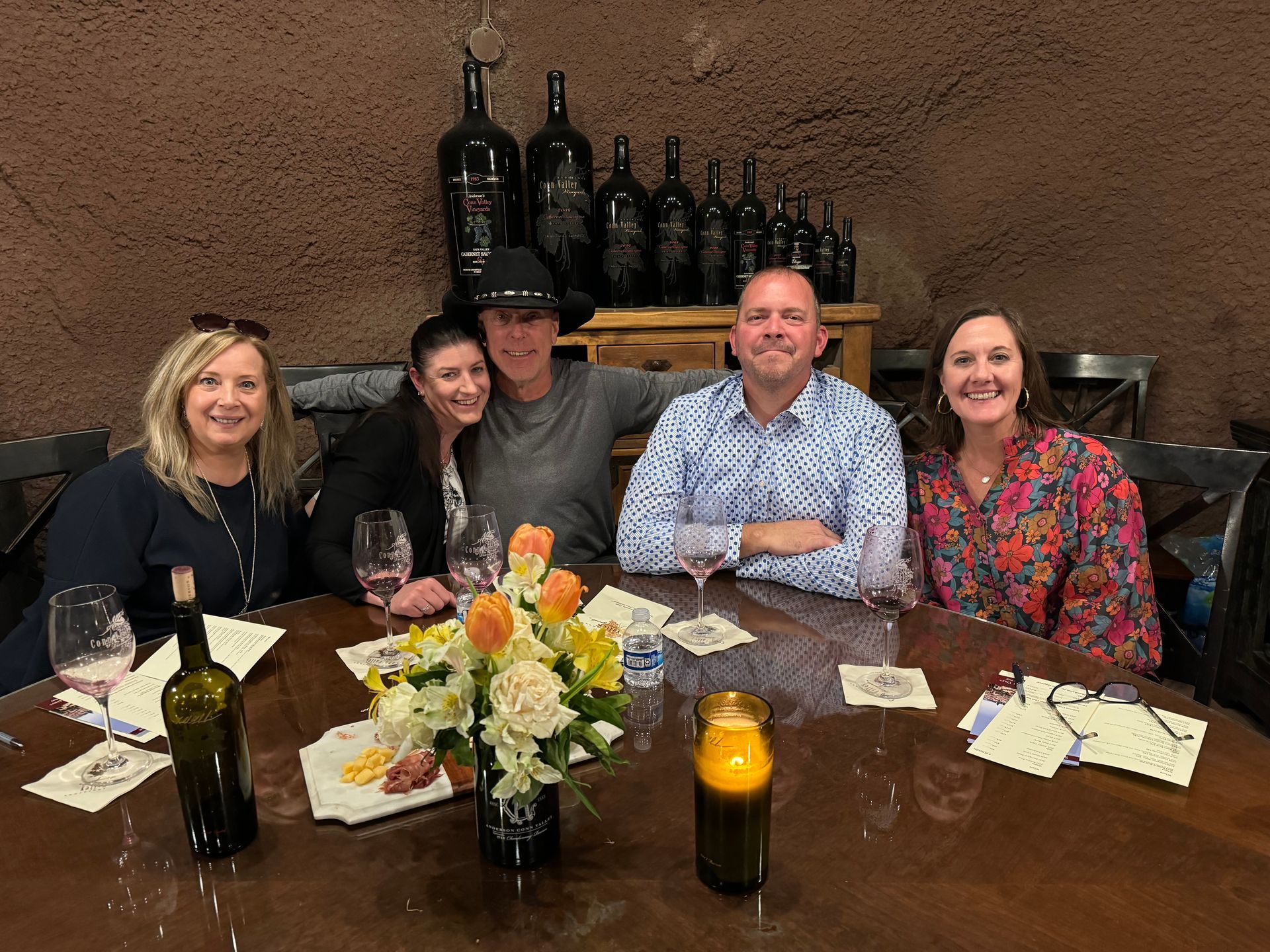 Five people smile at a round table with wine and flowers in a rustic, stone-walled cellar.