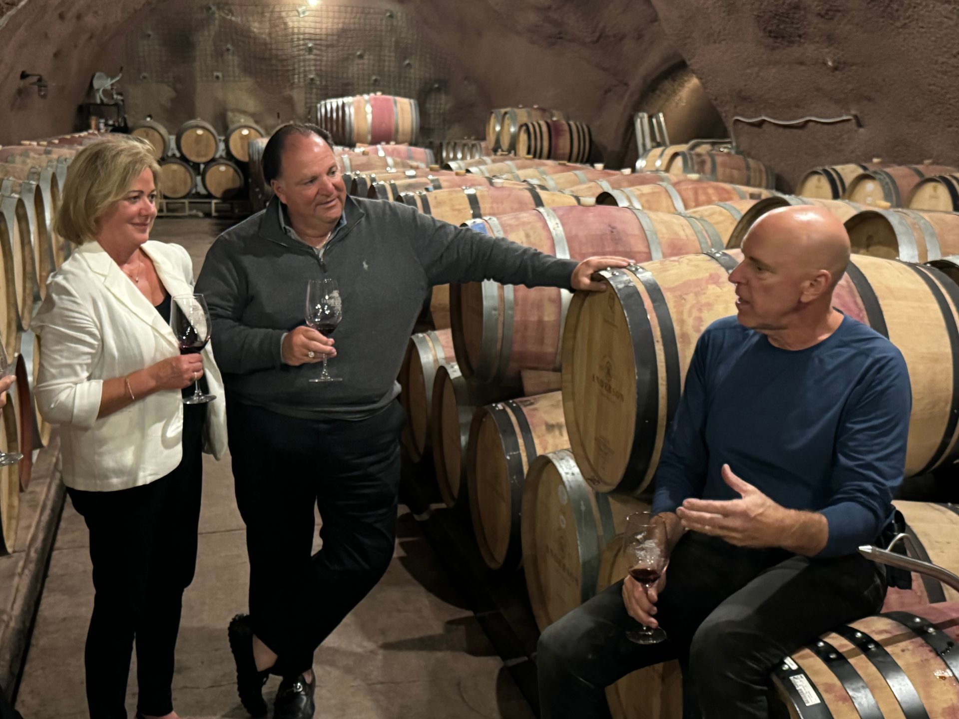 Three people hold wine glasses and chat in a wine cellar surrounded by stacked oak barrels.