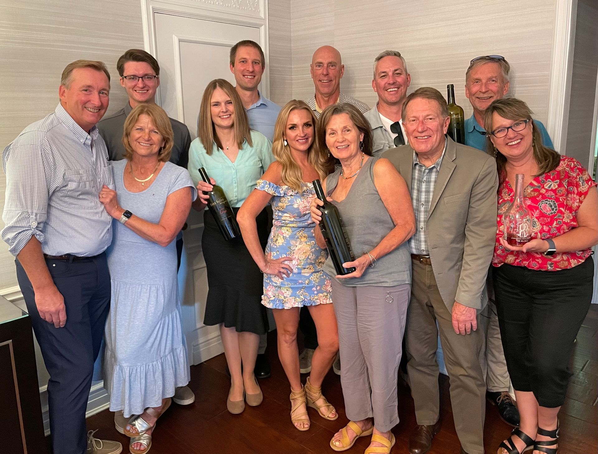 A group of twelve people stand together indoors, smiling while holding wine bottles and a carafe, posed for a photo.