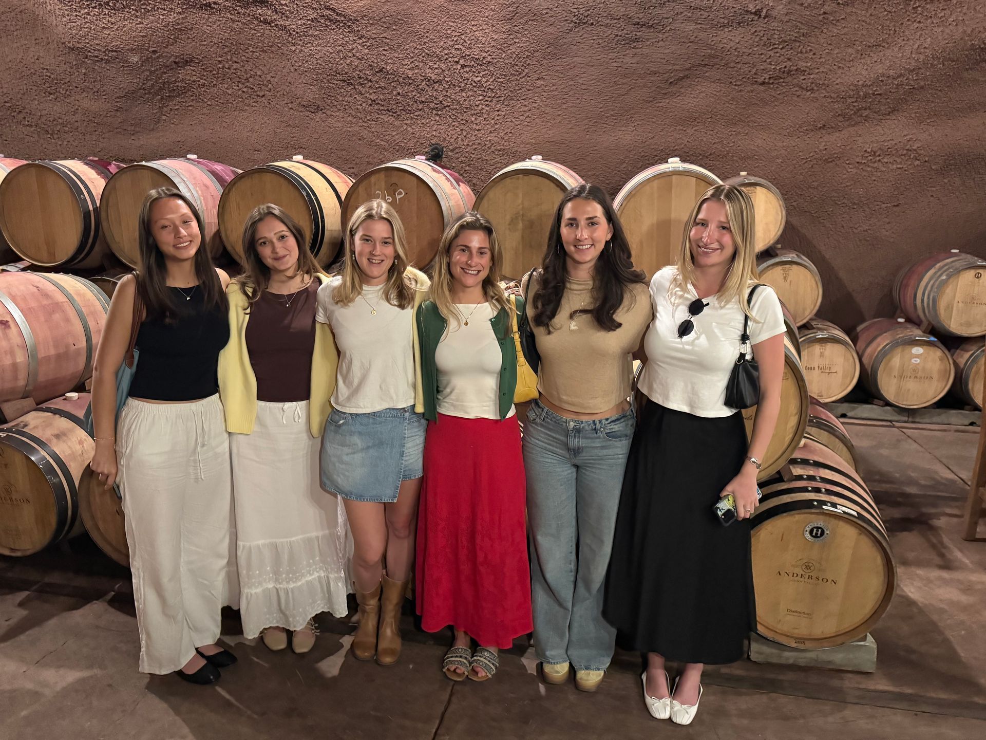 Six people stand in a line smiling in a wine cellar filled with wooden barrels, wearing casual tops, skirts, and jeans.