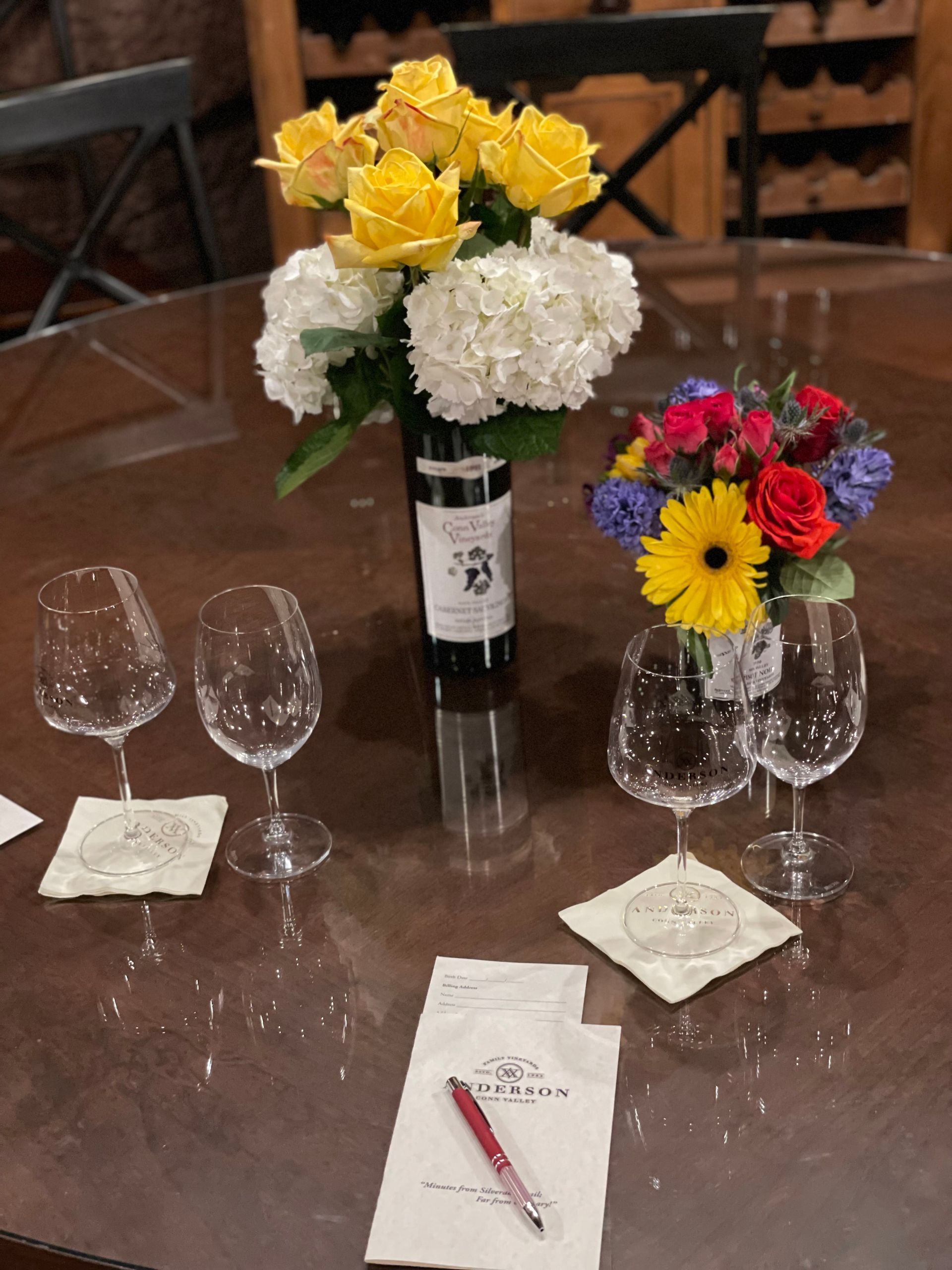 A wine tasting setup on a glass table with two bouquets of flowers, four empty wine glasses, and a pen on a napkin.