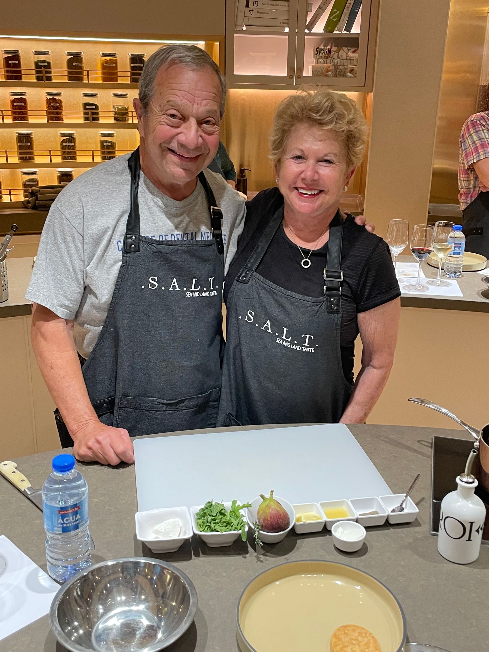 Two people in branded dark gray aprons smile while standing behind a counter prepared with ingredients for a cooking class.