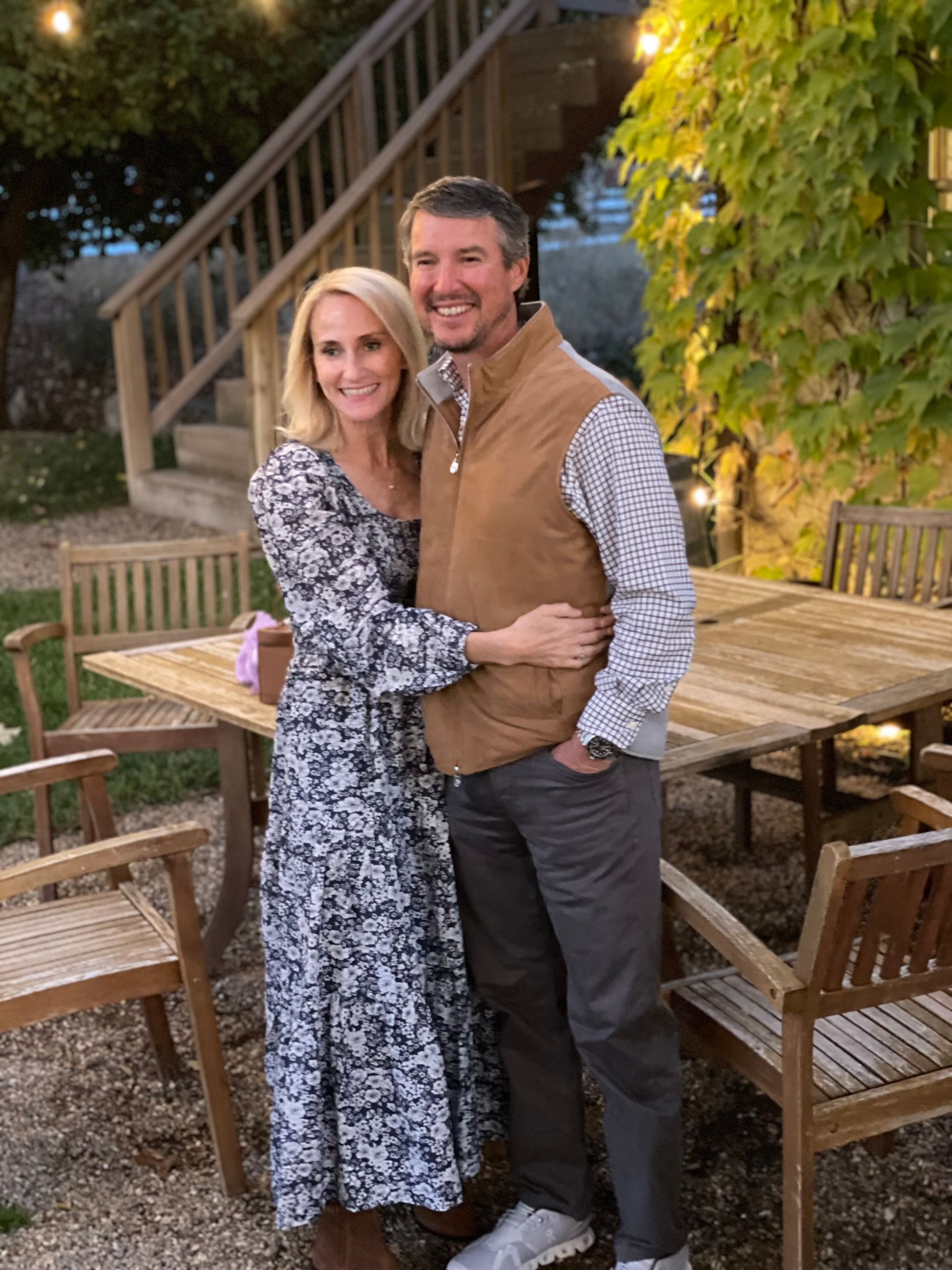 A smiling couple poses for a photo outdoors near a wooden table and stairs during twilight.