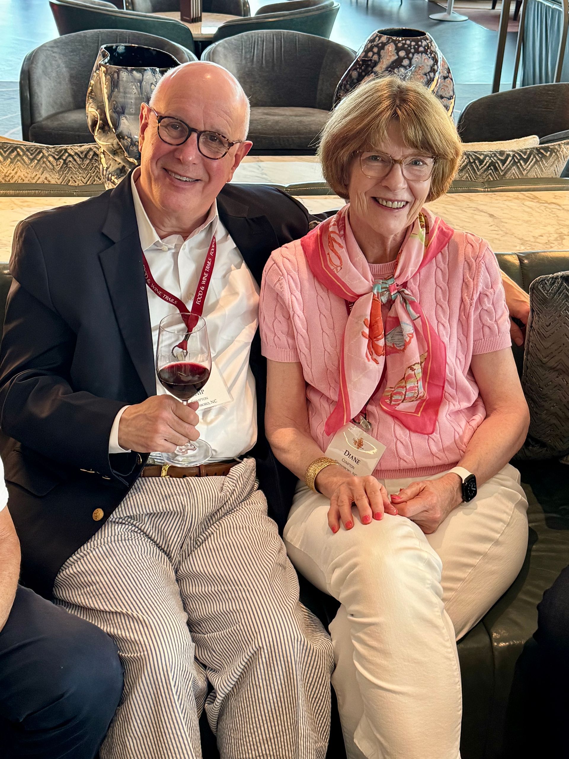 A couple smiles while sitting on a sofa in a lobby; the person on the left holds a wine glass, wearing a blazer and scarf.