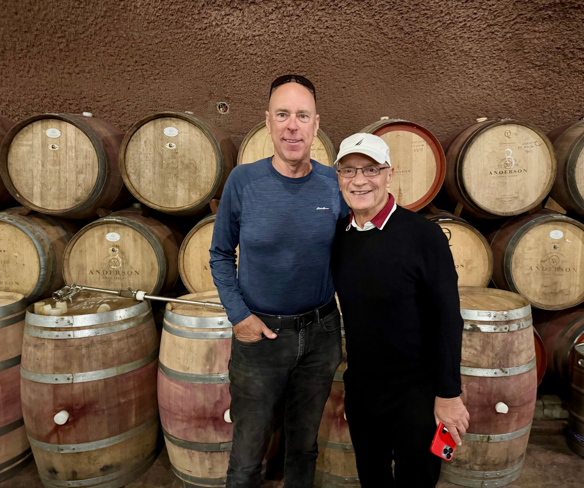 Two people stand smiling in a wine cellar surrounded by wooden barrels stacked against a textured wall.