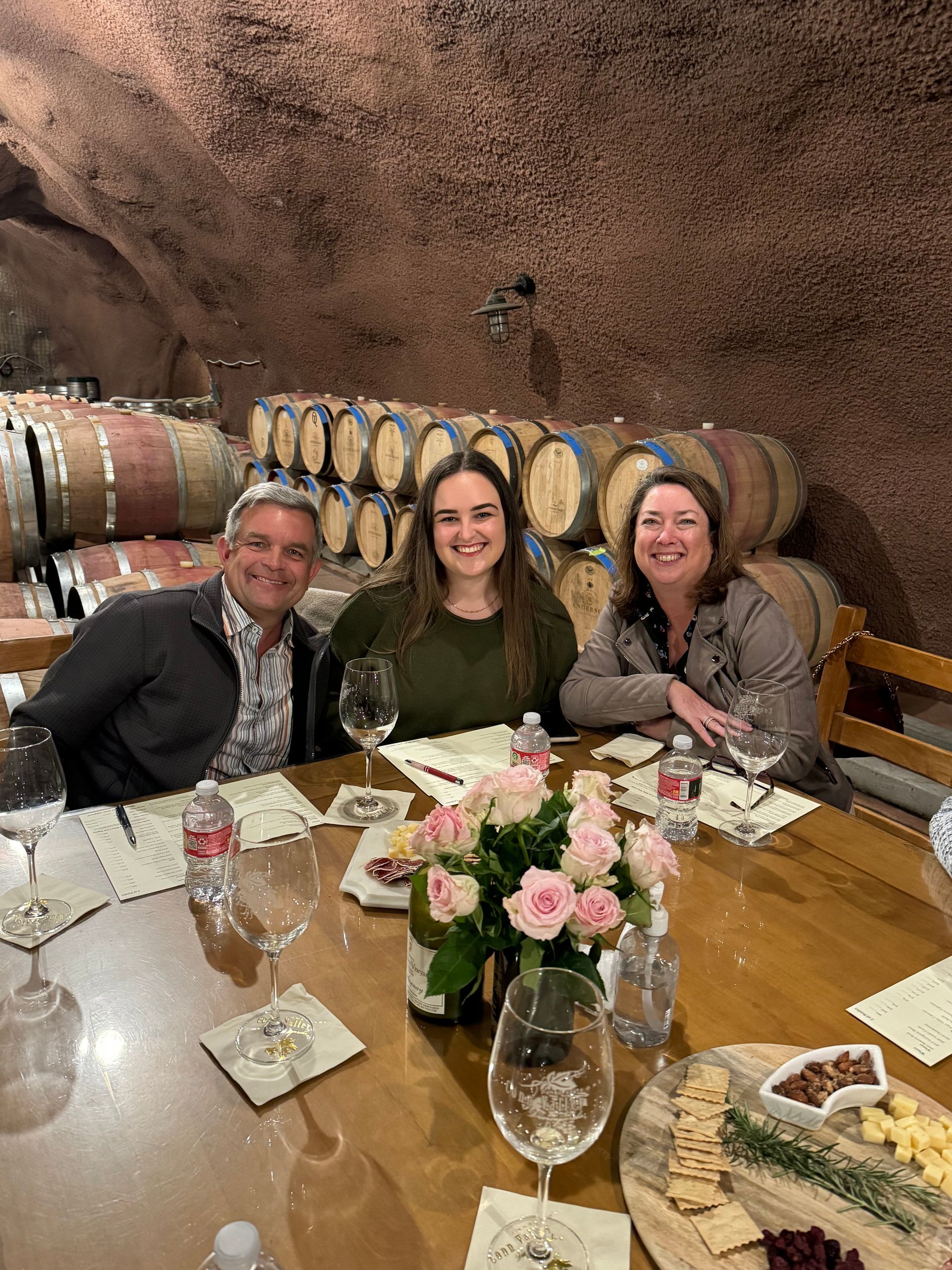 Three people smile at a table with wine, snacks, and a bouquet in a rustic wine cellar lined with wooden barrels.