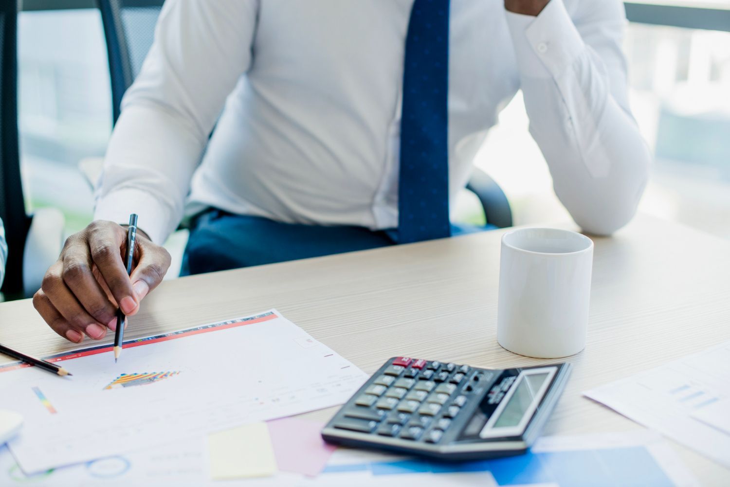 Man in white shirt and blue tie at a desk, holding a pencil and looking at papers with a calculator and mug.