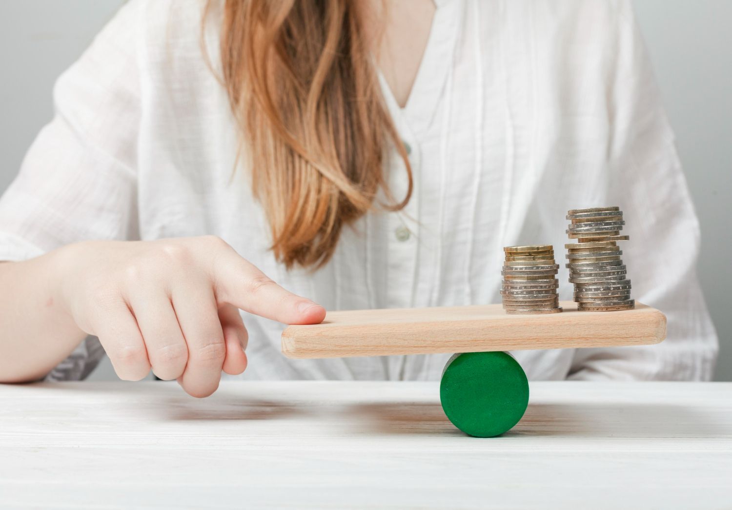 Woman's finger points to uneven stacks of coins on a wooden seesaw balanced on a green cylinder.