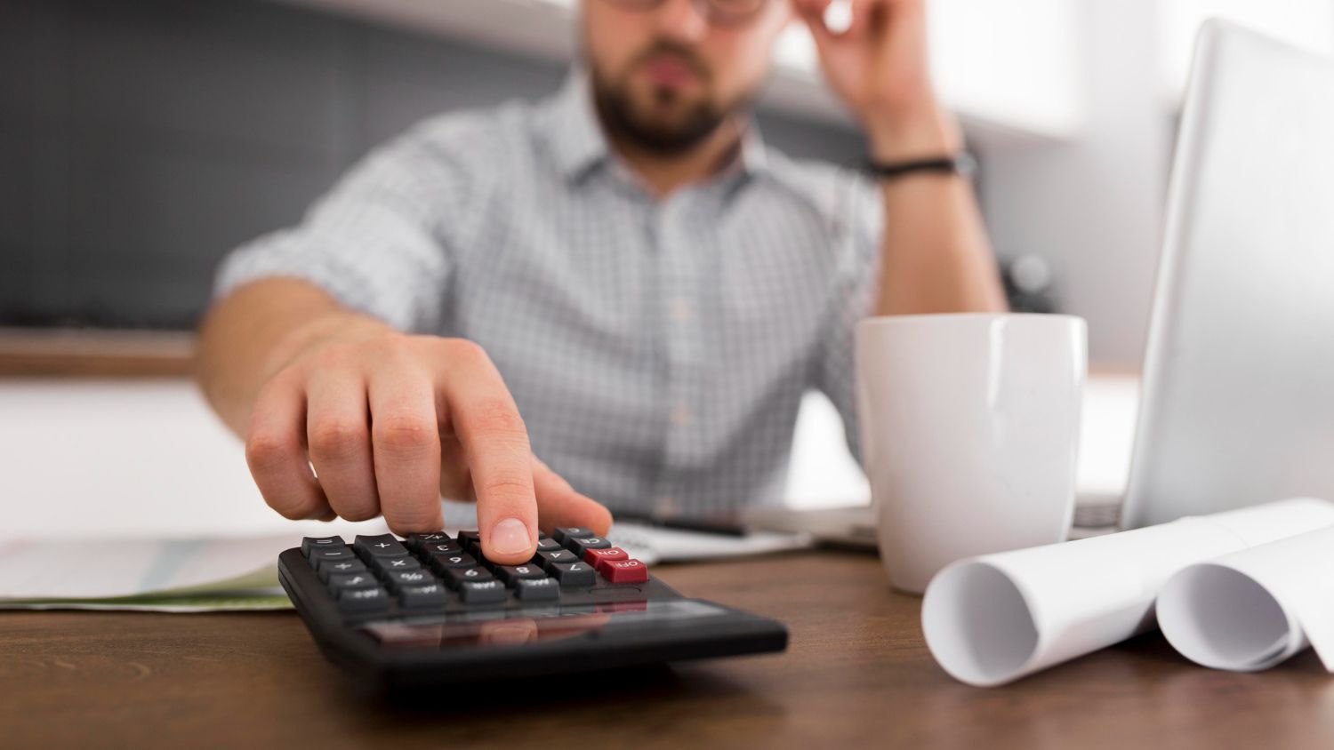 Man using a calculator at a desk, looking concerned, with papers, a laptop, and a mug.
